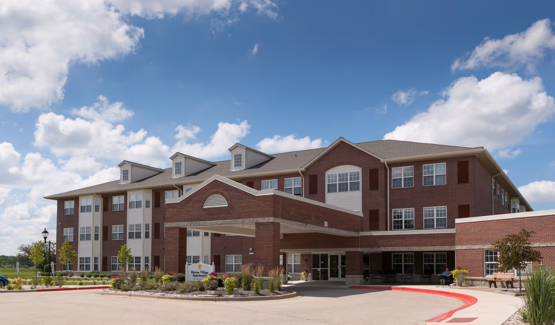 Exterior view of a three-story brick building with white trim and multiple windows under a blue sky with scattered clouds. The building has a covered entrance with a sign that reads Marian Village Apartments. There are some small trees and landscaped plants around the entrance and a curved driveway in front.