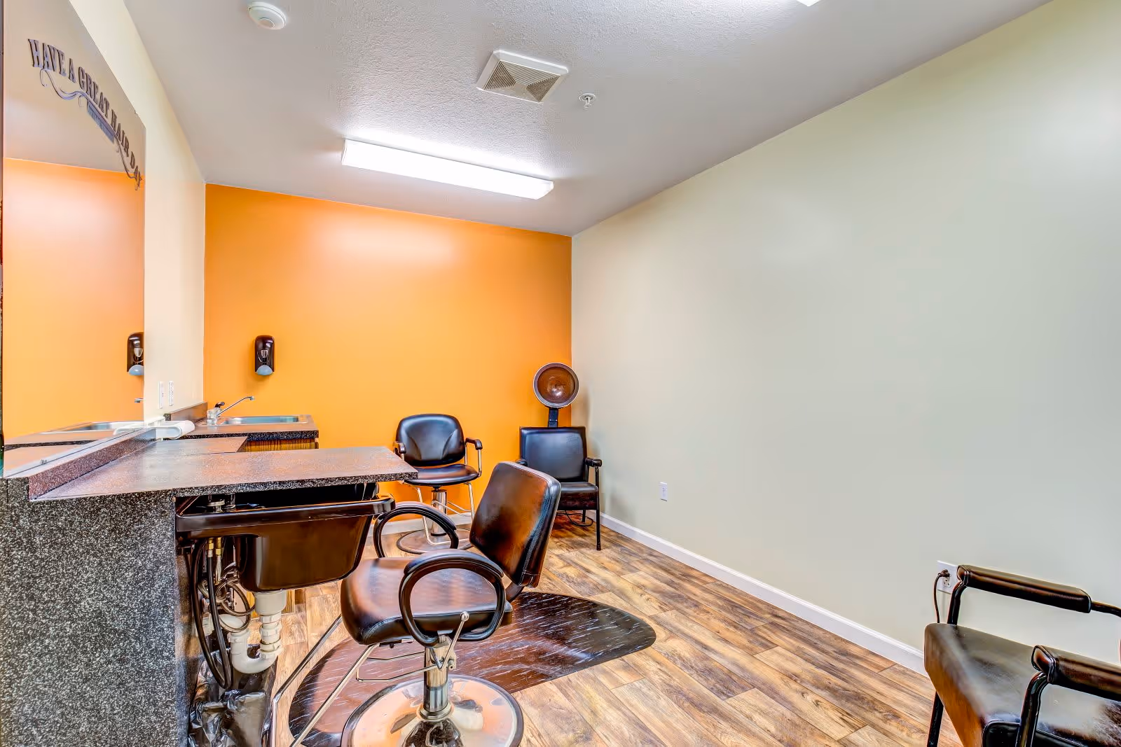 Interior view of a small salon or hair styling area with three black salon chairs, a hair washing sink, a large mirror, and an orange accent wall. The floor has wood-like vinyl flooring and the room is lit by a ceiling light.