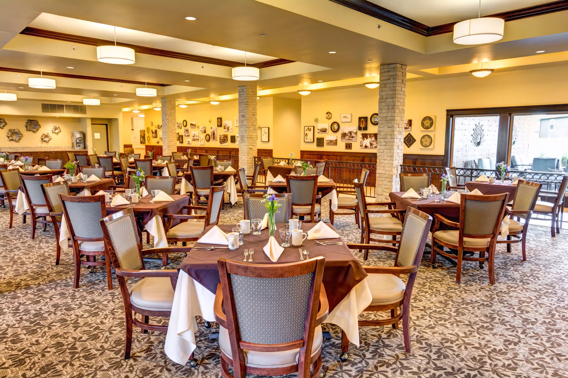 A spacious dining room with multiple tables covered in white and brown tablecloths, each set with napkins, cups, and silverware. The room has patterned carpet flooring, beige walls decorated with framed pictures and plates, and large windows letting in natural light. Ceiling lights provide additional illumination.