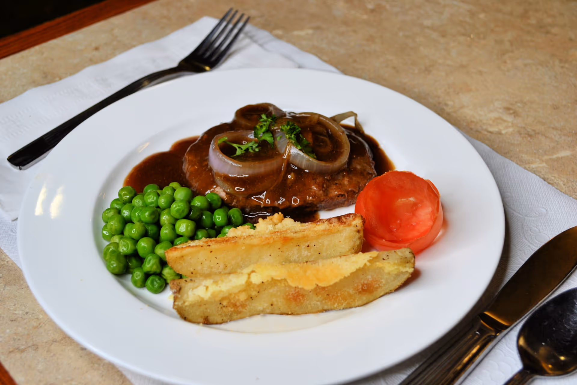 A white plate with a meal consisting of green peas, two potato wedges, a slice of tomato, and a hamburger patty topped with brown gravy and cooked onions, placed on a white napkin with a fork, knife, and spoon on a beige table surface.