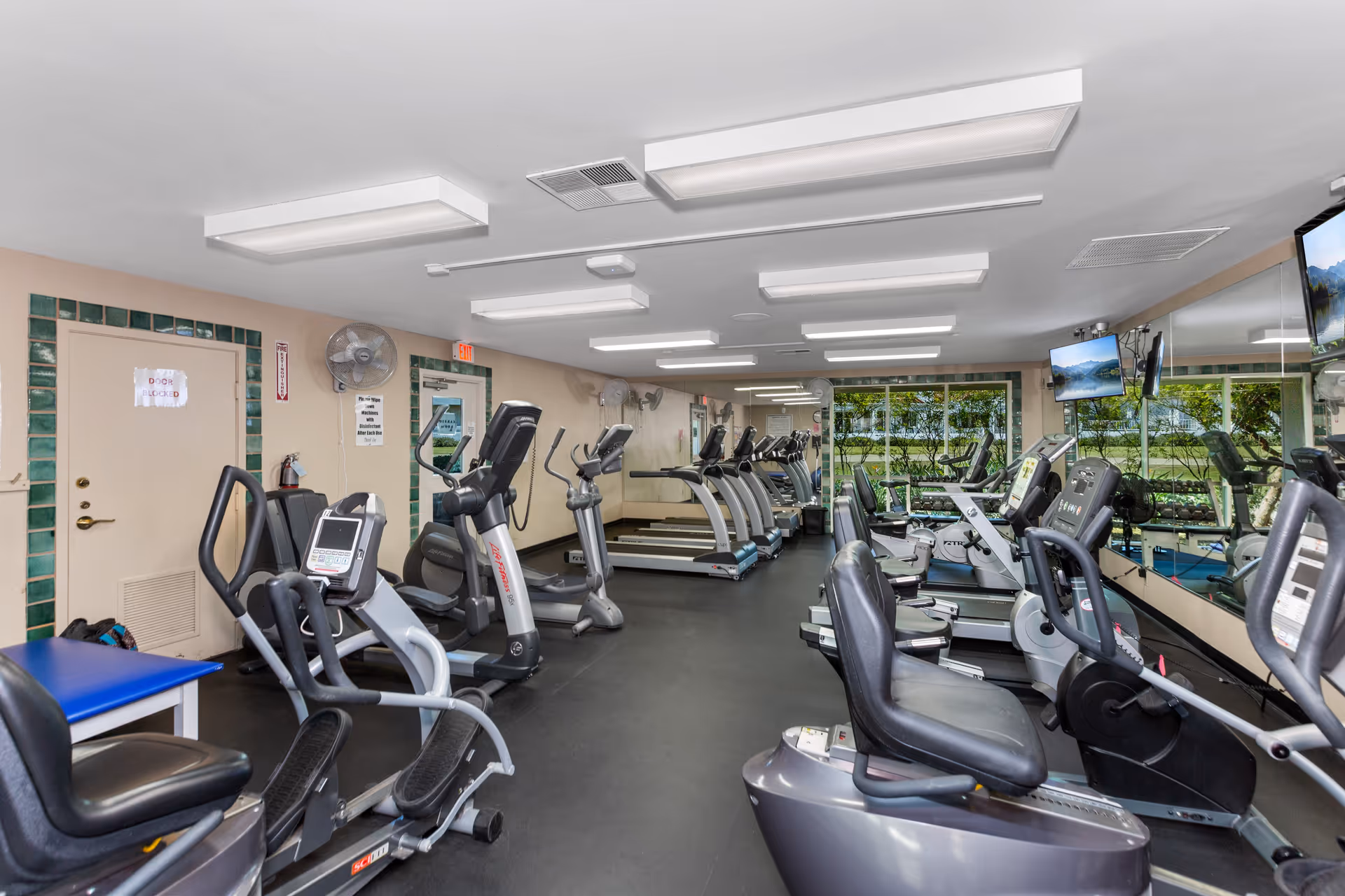 Interior view of a fitness room with multiple exercise machines including treadmills, stationary bikes, and elliptical machines. The room has bright overhead lighting, large mirrors on one wall, and windows showing greenery outside.