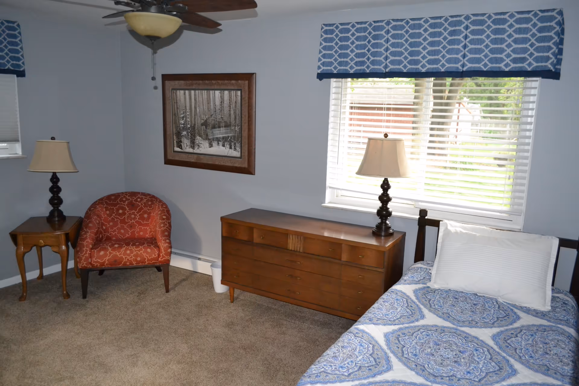 A bedroom with a single bed covered in a blue and white patterned bedspread and a white pillow. Next to the bed is a wooden dresser with a lamp on top. There is a window with white blinds and a blue patterned valance above it. In the corner of the room, there is a red patterned armchair next to a small wooden side table with another lamp. A framed picture of a forest scene hangs on the light blue wall. The floor is carpeted in beige.