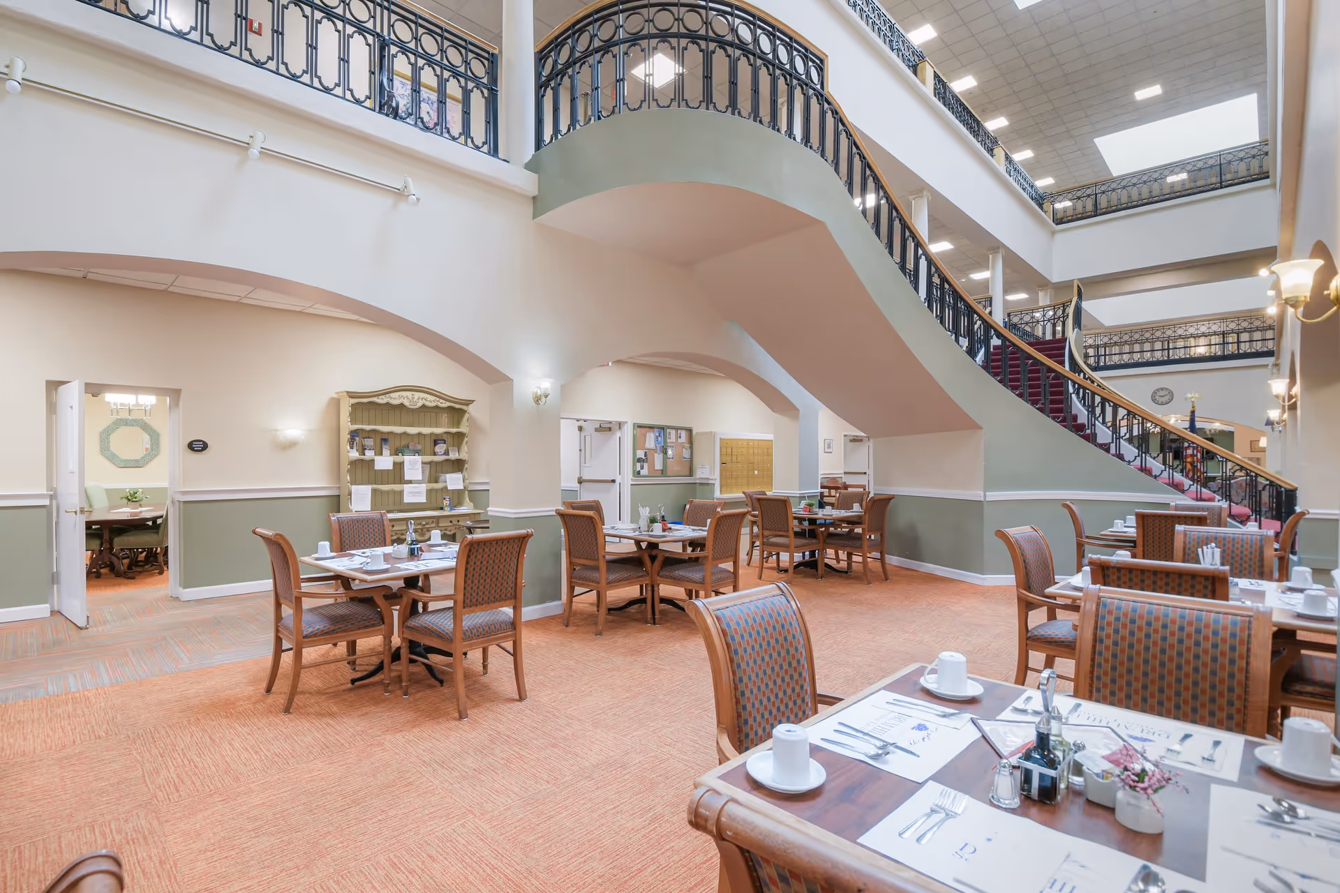 A spacious dining area in a senior living community with multiple wooden tables and chairs arranged neatly. The tables are set with white cups, plates, and silverware. The room features a high ceiling with a large staircase and decorative black railings. The walls are painted in a soft beige and green color, and there is a carpeted floor in a warm orange tone. In the background, there is a doorway leading to another room with a green chair and a decorative mirror on the wall.