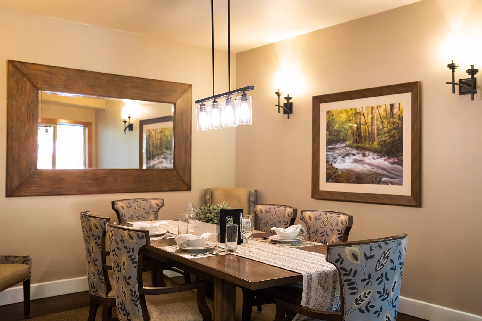 A dining room with a wooden table set for six people, featuring patterned upholstered chairs, a table runner, glassware, and white dishes. The room has warm lighting from wall sconces and a hanging light fixture. A large wooden-framed mirror and a framed picture of a forest stream decorate the beige walls.