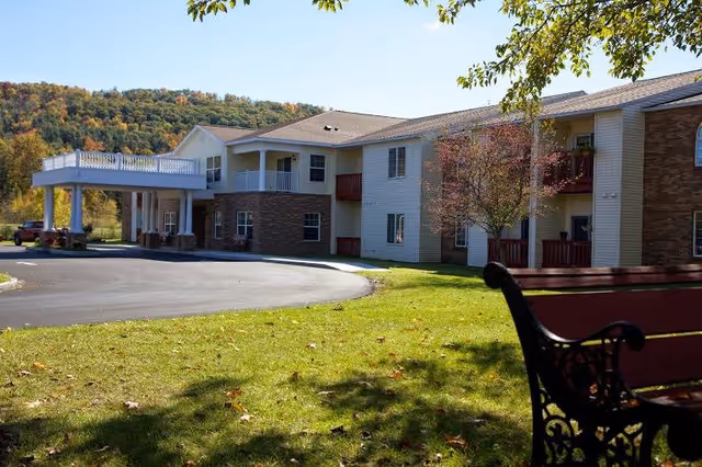 Exterior view of a two-story senior living facility building with a covered entrance, surrounded by green grass and trees with autumn foliage. A decorative bench is visible in the foreground on the right side.