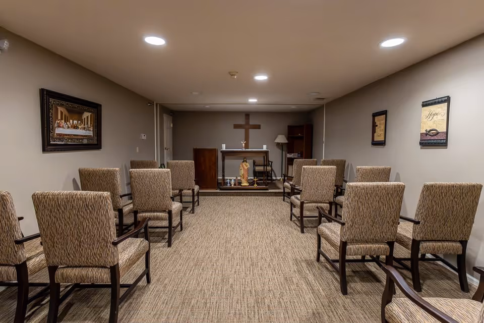 A small chapel or prayer room with beige walls and carpeted floor. There are two rows of patterned upholstered chairs facing a small altar with a wooden cross on the wall behind it. Religious statues and candles are placed on the altar. Framed religious artwork hangs on the walls.
