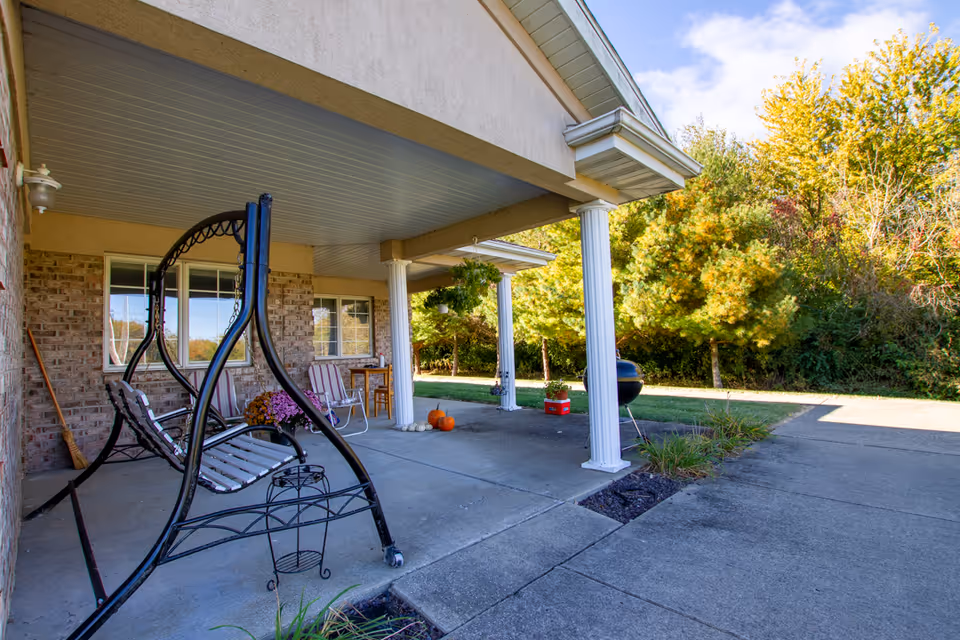 Covered outdoor patio area with a black metal swing, two striped chairs, a small table, pumpkins on the ground, and a charcoal grill. The patio is supported by white columns and overlooks a driveway and green trees in the background.