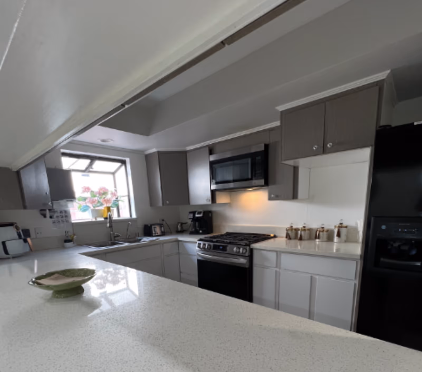 Modern kitchen with white countertops and gray upper cabinets. There is a stainless steel stove with a microwave above it, a black refrigerator, a coffee maker, a toaster, and a window with a flower pot on the windowsill. A green dish with a napkin is on the countertop in the foreground.