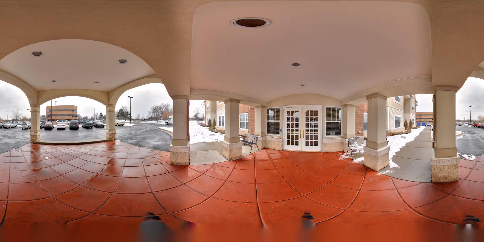 Covered entrance area of University Senior Living facility with red tiled floor, beige columns, and ceiling lights. There are benches on either side near the double glass doors leading inside. Outside, there is a parking lot with several cars and some snow on the ground and sidewalks.