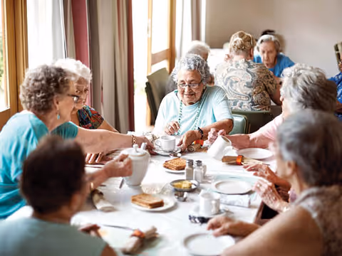 Several elderly residents seated around a dining table sharing tea, toast, and conversation in a bright communal dining room.
