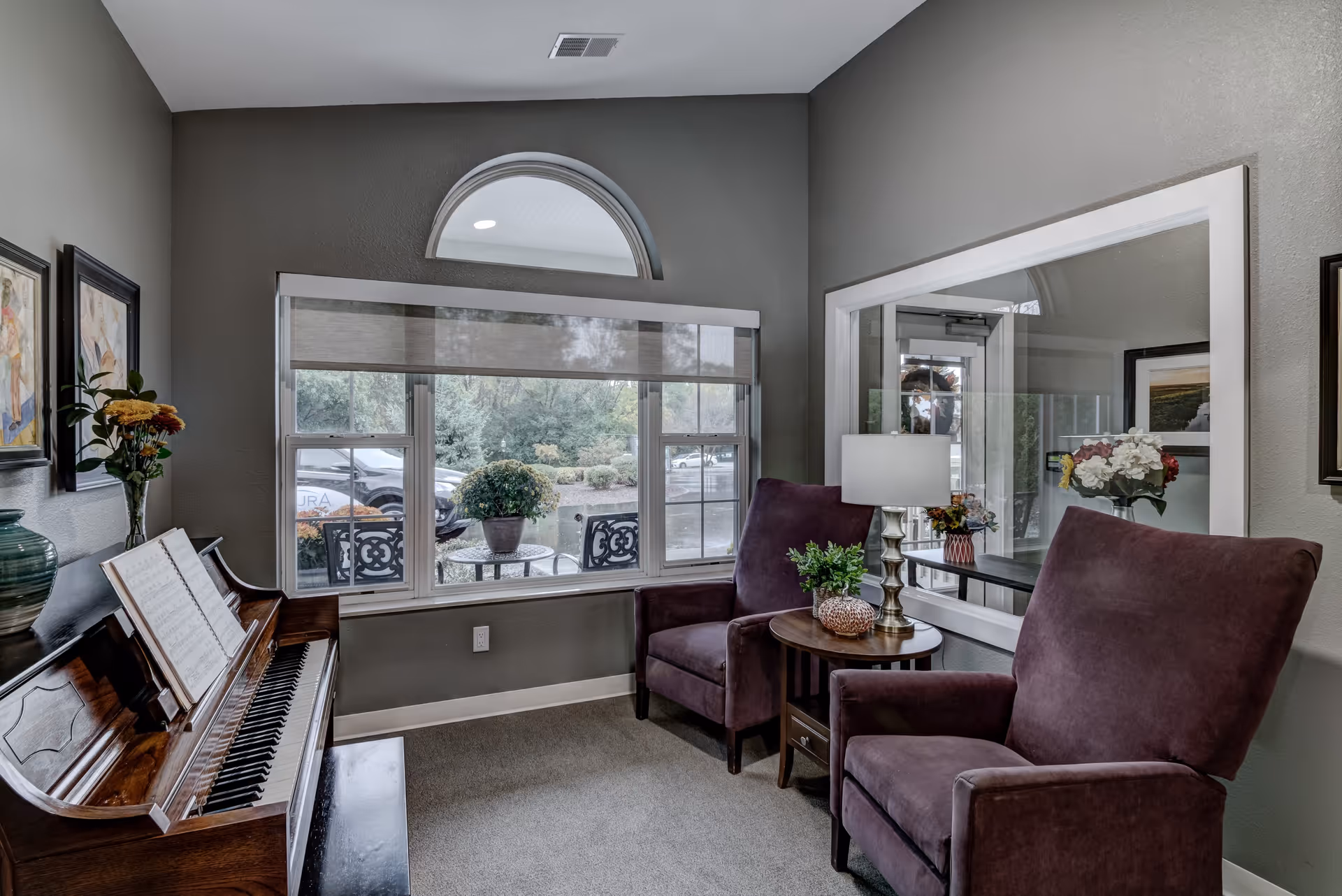 A cozy sitting area in a senior living facility with two purple armchairs separated by a small wooden side table holding a lamp and a small plant. To the left, there is an upright piano with sheet music and a vase of flowers on top. A large window with a half-circle window above it shows an outdoor patio with chairs and a table, and greenery beyond. The walls are painted gray and decorated with framed artwork.