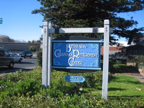 A blue sign with white text reading 'Bethesda Christian Retirement Center' mounted on white wooden posts, surrounded by green bushes and grass, with a parking lot and buildings in the background under a clear sky.