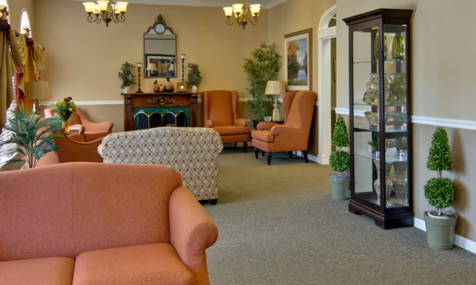 Comfortable senior living lounge with upholstered sofas and armchairs, decorative plants, a fireplace and a glass display cabinet.