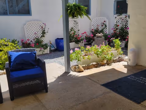 Outdoor patio area with a black wicker chair featuring a blue cushion, a garden bed with various flowering plants, white lattice panels against a white wall, and a hanging fern plant. There is a dark mat on the ground with partially visible text.