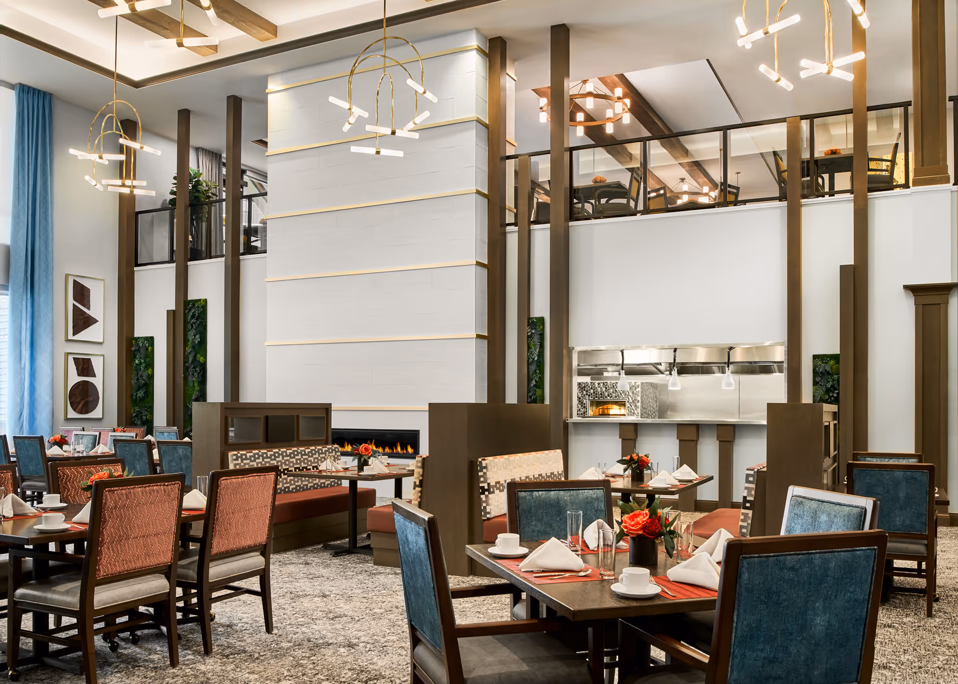 A spacious and elegant dining room in a senior living facility with tables set for meals, featuring chairs with blue and red upholstery, a modern fireplace, and contemporary light fixtures hanging from a high ceiling. There is a mezzanine level with additional seating and a view into a kitchen area with a pizza oven.
