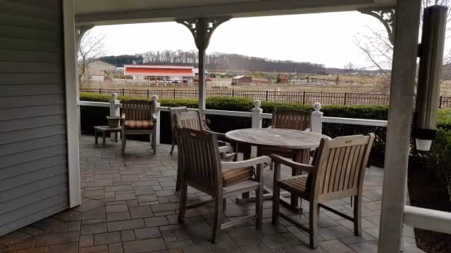 Covered outdoor patio area with a round wooden table and several wooden chairs on a tiled floor. Beyond the patio, there is a view of a fenced grassy area with a red barn and other farm buildings in the distance under a cloudy sky.