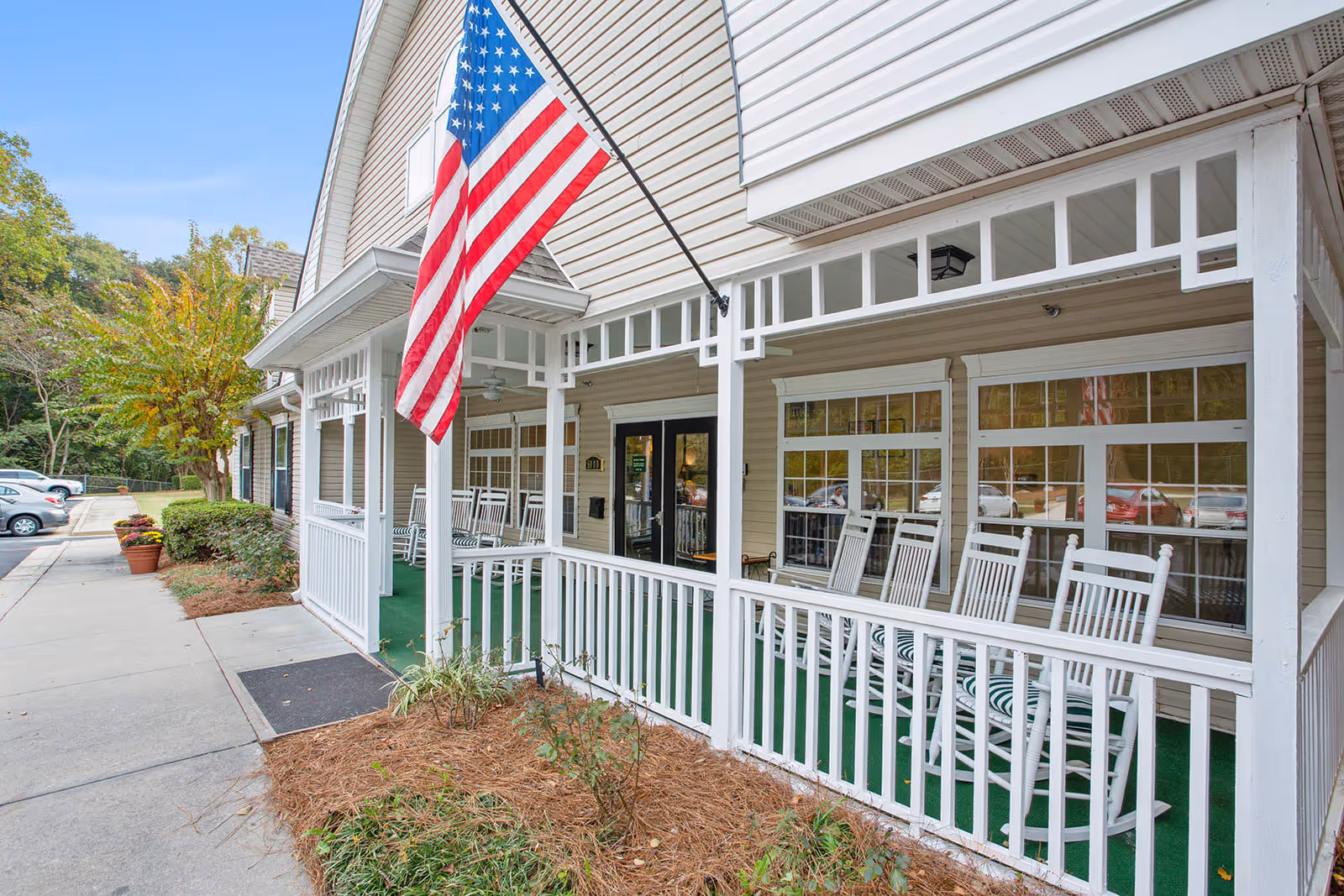 Front porch of a senior living facility with white rocking chairs lined up along a white railing, an American flag hanging from a pole, and a parking lot with cars in the background.