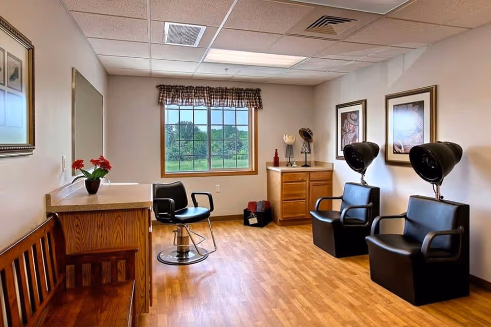 A bright room with wooden flooring and beige walls featuring a large window with a checkered valance. The room contains two black salon hair dryer chairs, a black salon styling chair, a wooden bench, a wooden cabinet with decorative items, and framed artwork on the walls.