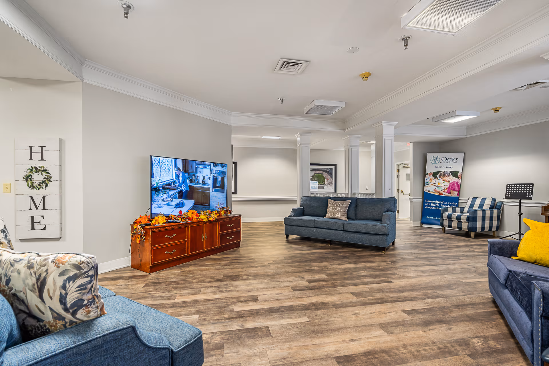 A spacious senior living facility common area with wood flooring and neutral walls. The room features multiple blue upholstered sofas and armchairs with decorative pillows. A wooden TV stand holds a flat-screen television displaying a cooking show. There is a decorative sign on the wall that spells HOME with a wreath in place of the letter O. In the background, a banner for Oaks at Cedar Shoals Senior Living is visible along with a music stand and piano.