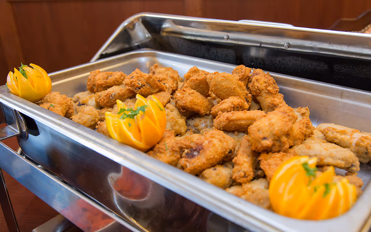 A stainless-steel chafing dish filled with breaded fried chicken pieces and carved orange garnishes.