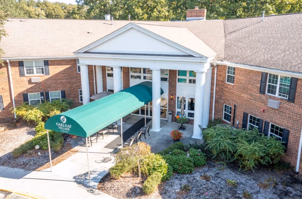 Front entrance of a brick senior living building with white columns and a green canopy reading "Oakleaf Village" over the walkway.