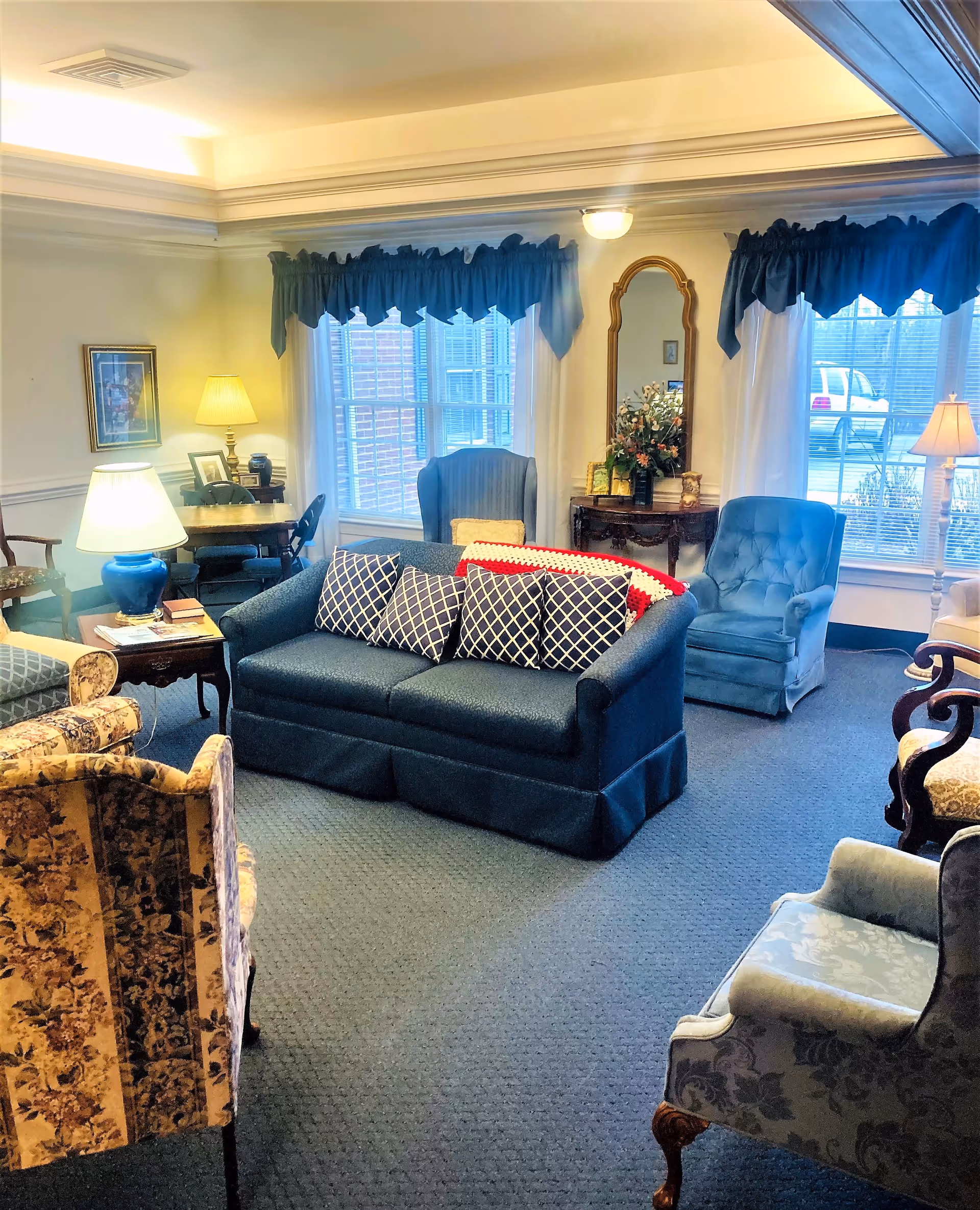 A cozy living room in a retirement community with a blue sofa adorned with patterned pillows, several upholstered armchairs in various floral and solid fabrics, a wooden side table with a lamp, and large windows with blue valances letting in natural light. There is also a small table with a mirror and floral arrangement against the wall.
