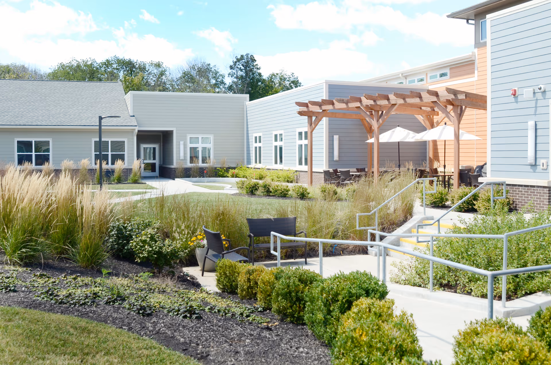 Outdoor courtyard area at Wellbrooke of Avon featuring a pathway with handrails, landscaped greenery, ornamental grasses, a wooden pergola with tables and umbrellas, and light-colored building exteriors under a partly cloudy sky.