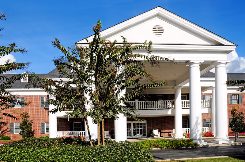 Front entrance of a red-brick building with large white columns, a covered portico and balcony surrounded by landscaping.