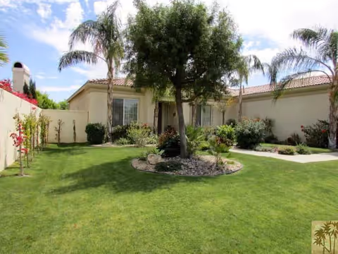 Front courtyard of a single-story residential building with a green lawn, palm trees, and landscaped plantings.