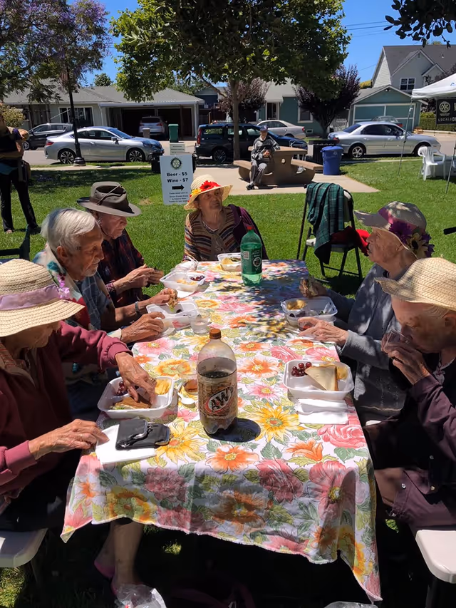 A group of elderly people sitting around a picnic table outdoors on a sunny day, eating food from takeout containers. The table is covered with a colorful floral tablecloth. Some of the people are wearing hats. In the background, there are cars parked on the street and a person sitting on a bench. A sign nearby indicates prices for beer and wine.