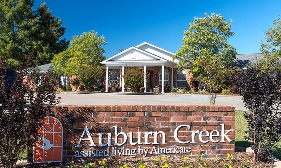 Exterior view of Auburn Creek assisted living facility showing a brick sign with the facility name in front of a single-story building with a white porch, surrounded by trees and greenery under a clear blue sky.