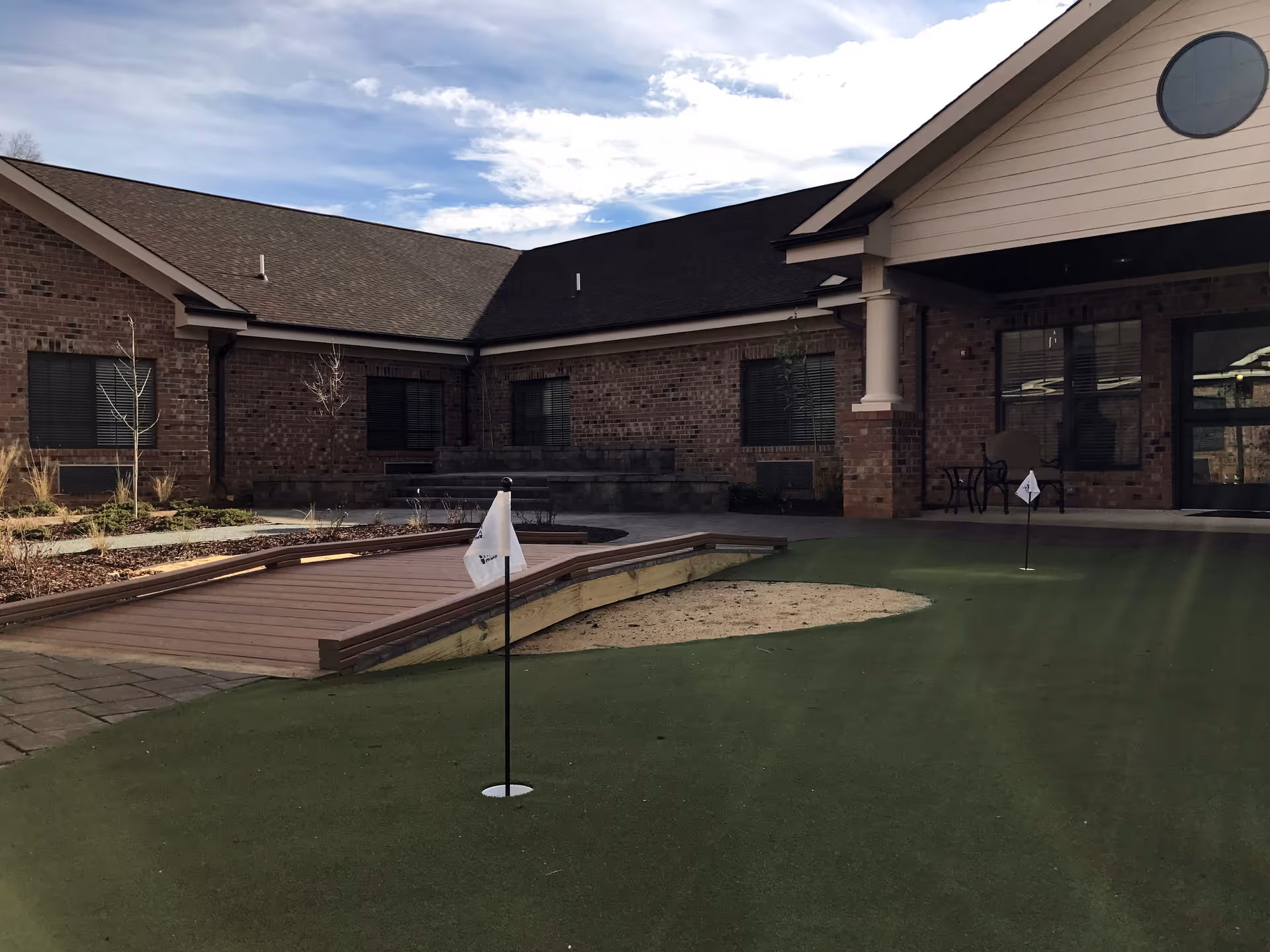 Brick senior living facility courtyard with a small putting green, practice flags, a wooden walkway and a covered entrance.