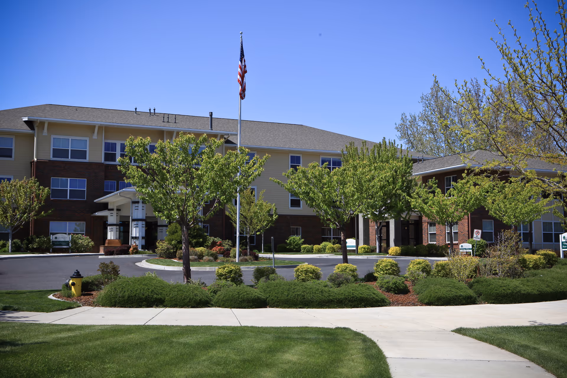 Front exterior of a three-story senior living building with a flagpole, landscaped circular driveway, and trees.