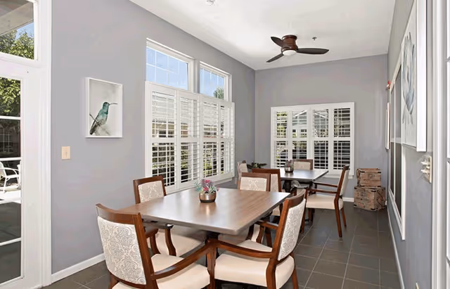 A bright dining room with two wooden tables, each surrounded by four upholstered chairs. The room has large windows with white plantation shutters allowing natural light to fill the space. There is a ceiling fan with wooden blades, light gray walls adorned with framed artwork, and a tiled floor. A door on the left leads to an outdoor area with patio furniture.