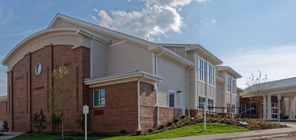 Exterior view of a senior living facility building with brick and light-colored siding, large windows, a curved roof section, and a landscaped area with grass and small trees under a partly cloudy sky.