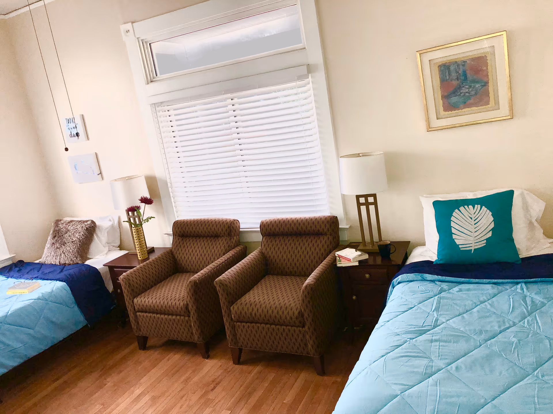 A bright room with two single beds on either side, each with blue bedding and decorative pillows. Between the beds are two brown patterned armchairs and two wooden nightstands with lamps. A window with white blinds is centered on the back wall, and a framed painting hangs above the bed on the right.