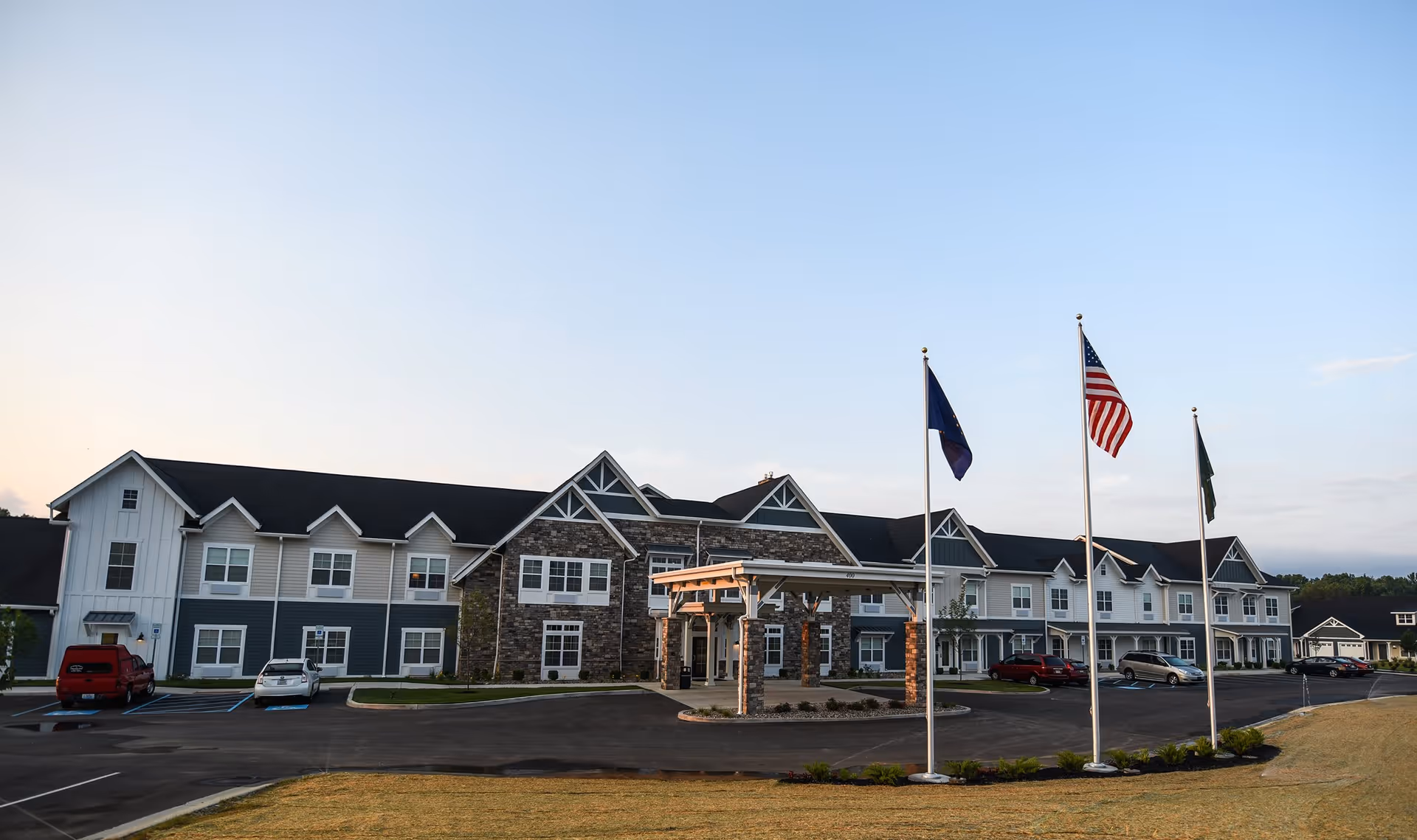 Exterior view of a two-story senior living facility building with a covered entrance, multiple windows, and a parking lot with several cars. Three flagpoles with flags are positioned in front of the building under a clear sky.