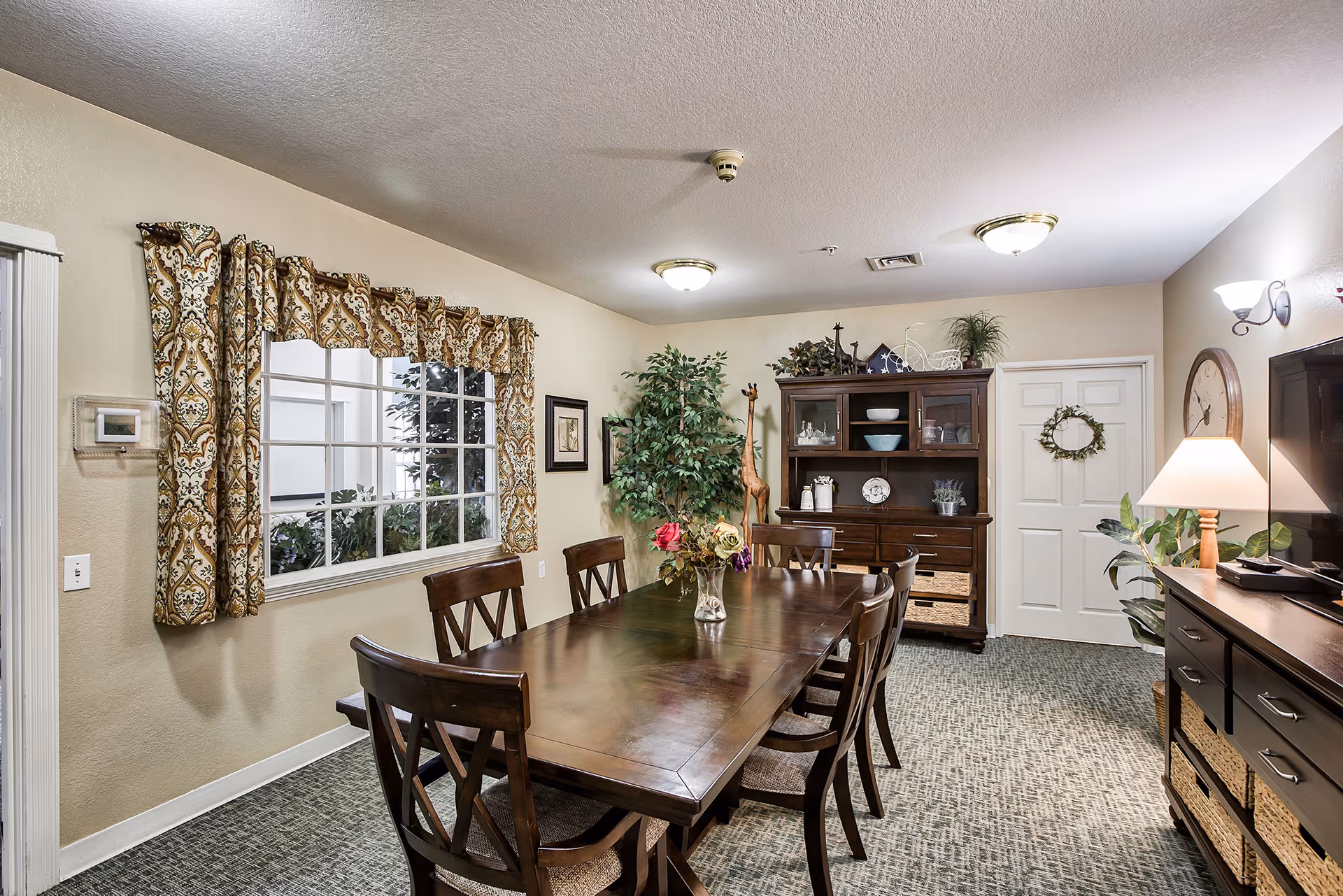 A well-lit dining room with a long wooden table surrounded by eight matching wooden chairs. The table has a vase with colorful flowers in the center. There is a large window with patterned curtains on the left wall, and a wooden cabinet with decorative items and plants against the back wall. A tall artificial plant stands in the corner, and a door with a small wreath is visible. The room has carpeted flooring and warm beige walls, with ceiling lights and a table lamp providing illumination.