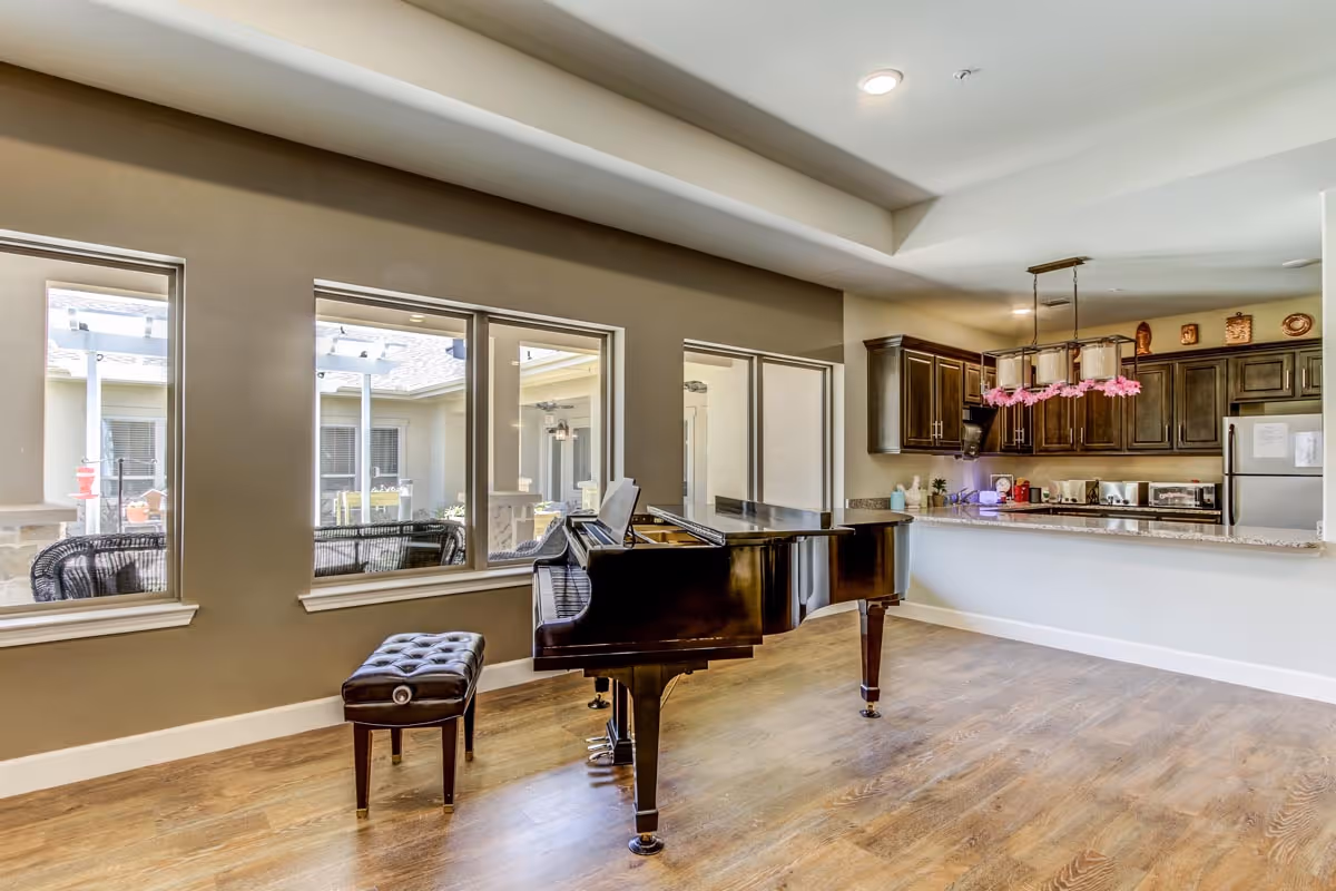 Interior view of a senior living facility showing a black grand piano with a matching bench on a wooden floor. Behind the piano is a kitchen area with dark wood cabinets, a granite countertop, and stainless steel appliances. Large windows on the left side provide a view of an outdoor patio area with wicker furniture.