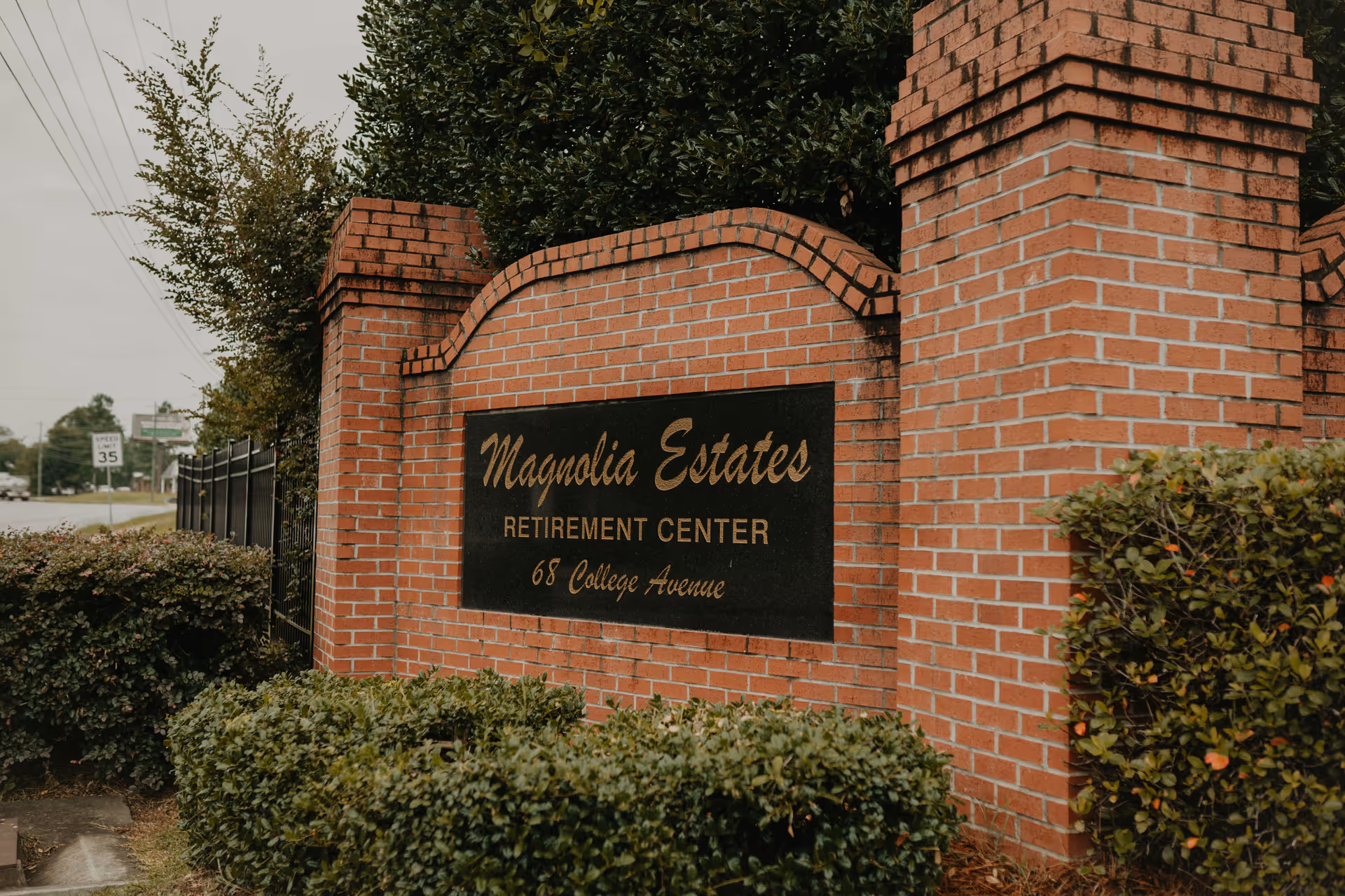 Brick entrance sign for Magnolia Estates Retirement Center at 68 College Avenue, surrounded by bushes and trees with a road and speed limit sign visible in the background.