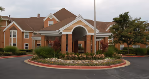 Front exterior view of a senior living facility named Brookdale Montrose featuring a covered entrance with brick pillars, landscaped roundabout with flowers and shrubs, and a multi-story building with brown roof and beige walls in the background.