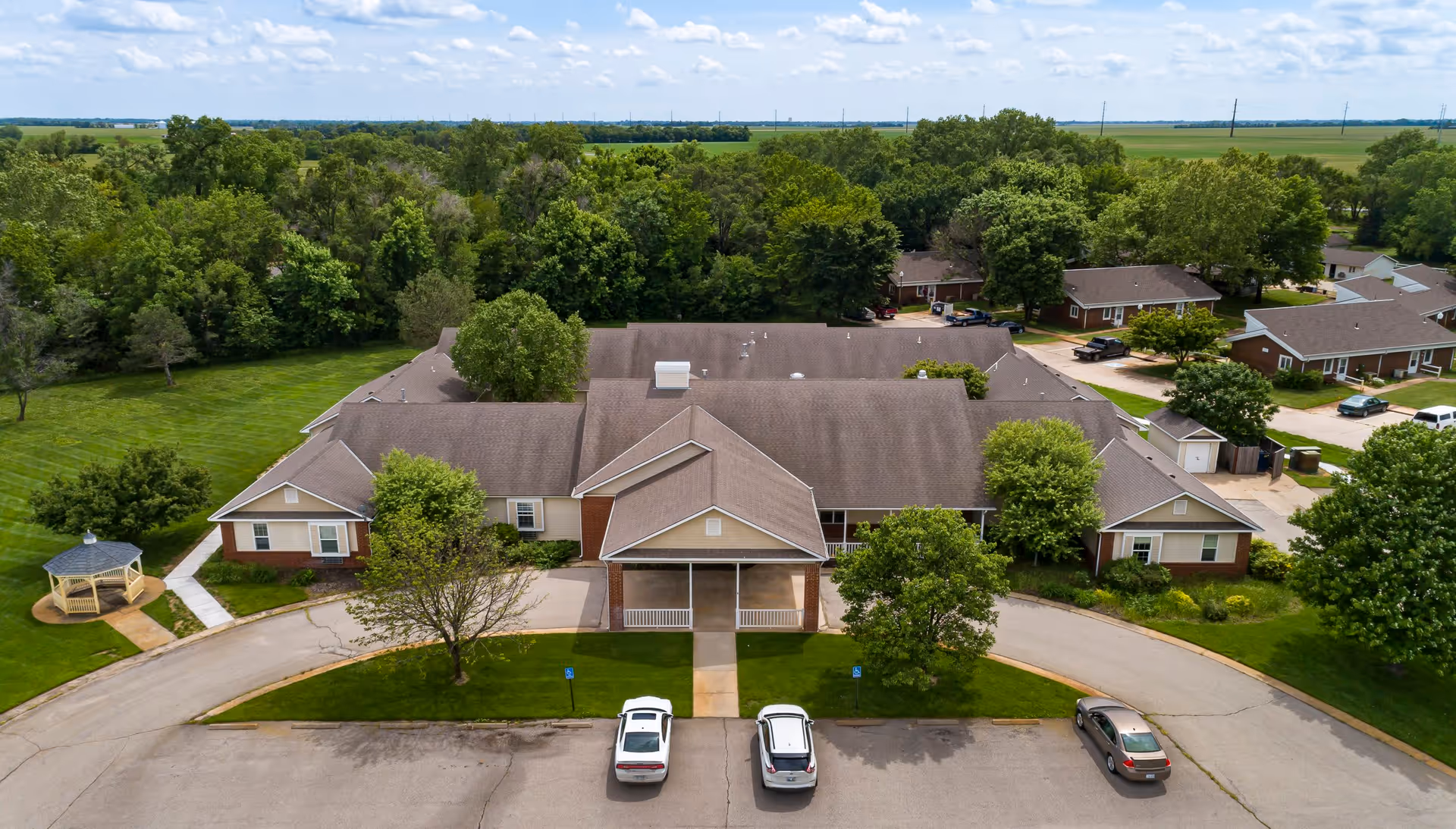 Aerial view of Homestead Assisted Living of Halstead, showing a single-story building with a large roof, surrounded by green trees and lawns. There is a circular driveway with three parked cars in front of the building, and a small gazebo is visible on the left side. The background features expansive farmland and a partly cloudy sky.