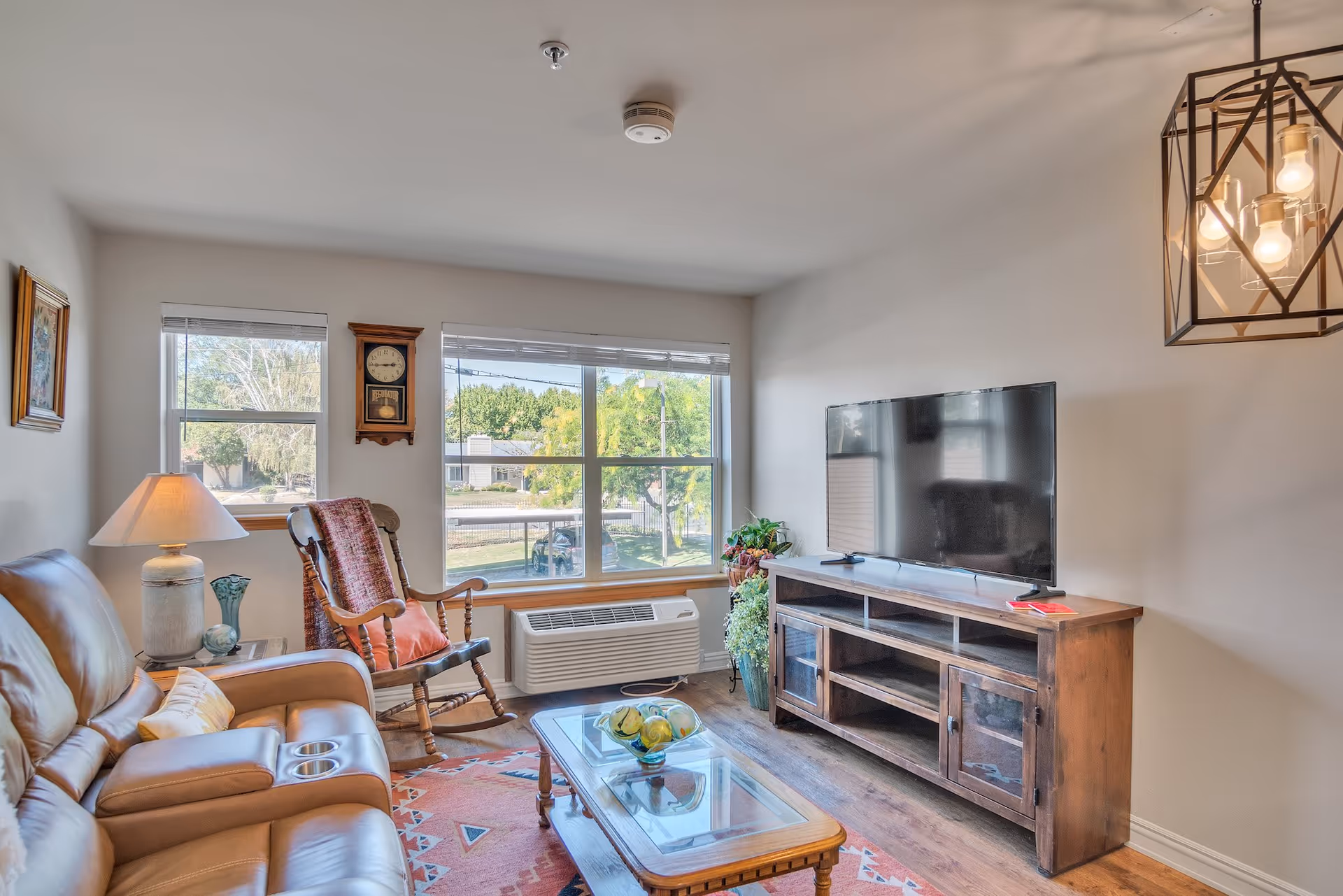 A cozy living room with a large window letting in natural light. The room features a brown leather sofa with cup holders, a wooden rocking chair with a cushion and throw, a wooden coffee table with a glass top and decorative bowl, a wooden TV stand with a flat-screen TV, a table lamp, a wall clock, and some potted plants. The floor has a patterned rug and the walls are painted light gray.