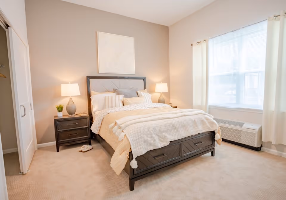 A bright and cozy bedroom with a large bed featuring beige and white bedding, two bedside tables with lamps, a small plant, slippers on the carpeted floor, a window with white curtains, and a closet with sliding doors.