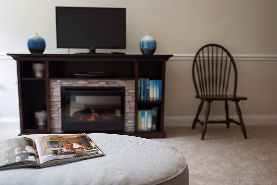 Living room with a television and electric fireplace in a dark wood console, a wooden chair against the wall, and an open book on a nearby ottoman.
