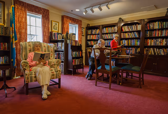 A cozy library room with red carpet and wooden bookshelves filled with books. An elderly woman in a pink sweater sits in a floral armchair reading a book. Two elderly people, a man and a woman, are standing and sitting near a wooden table also reading books. The room has two windows with red curtains and framed pictures on the walls.