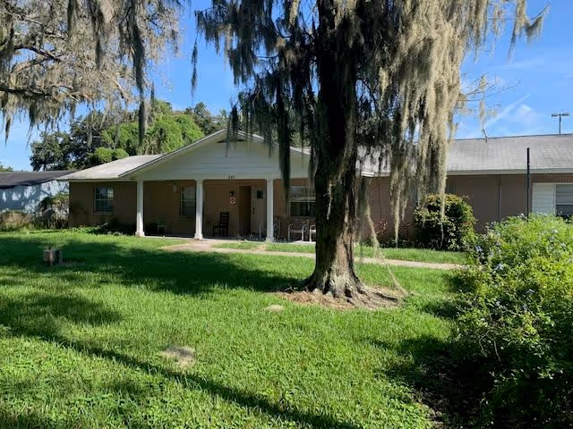 Single-story brick building with a covered front porch, green lawn, and a large moss-draped tree in front.