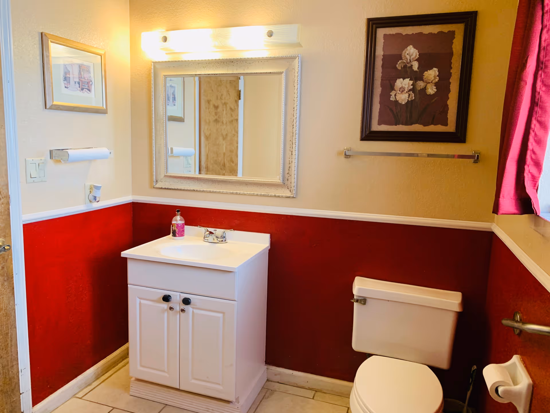 Small bathroom with a white vanity and sink beneath a framed mirror, a toilet, red lower walls and framed artwork.