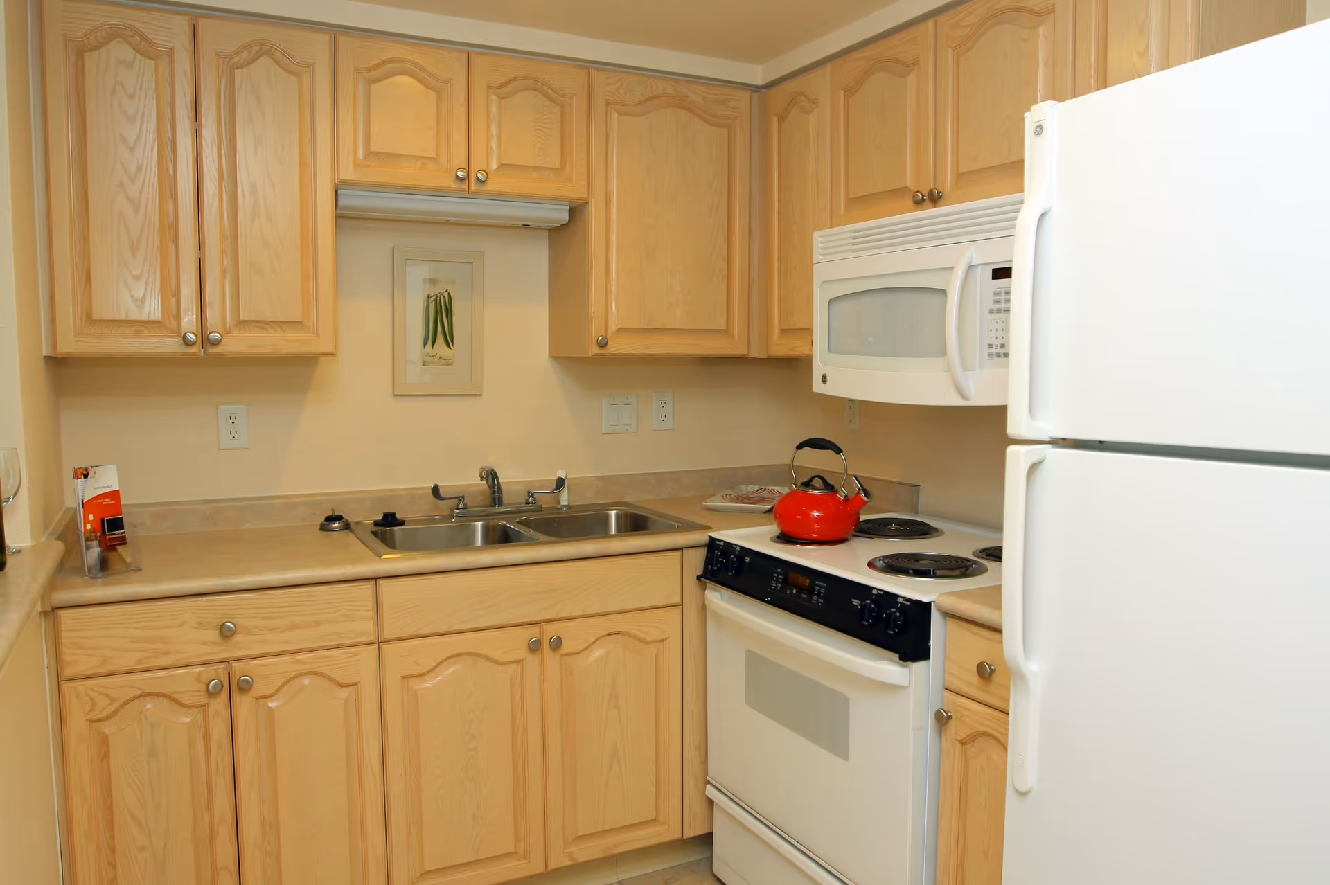 A compact kitchen with light wood cabinets, a double stainless steel sink, a white electric stove with a red kettle on top, a white microwave above the stove, and a white refrigerator. There is a small framed picture on the wall above the sink.