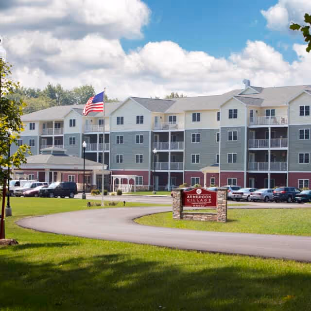 Exterior view of a multi-story senior living facility named Armbrook Village with a curved driveway, parked cars, an American flag on a flagpole, and a sign in front of the building. The building has balconies and a mix of light gray, white, and red siding under a partly cloudy sky.